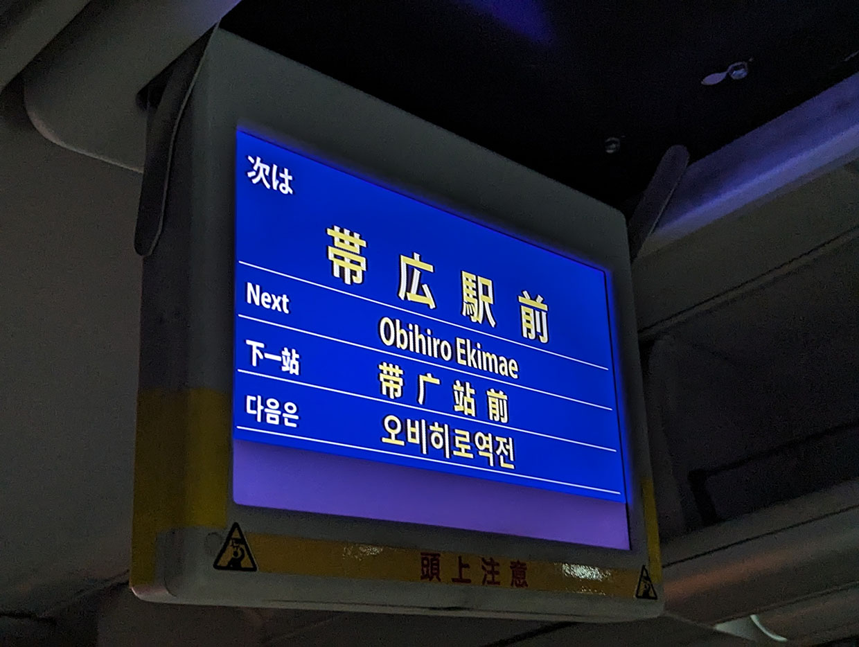 バス車内のモニターに表示された「次は 帯広駅前」の案内表示。日本語、英語、中国語、韓国語で表示されている。