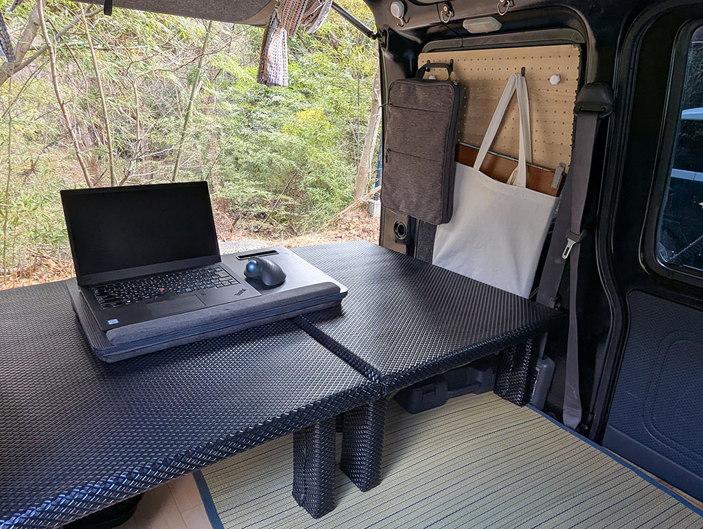軽バンの車内に設置されたワークスペース。黒のテーブル上にノートパソコンとトラックボールマウスが置かれており、窓の外には自然の風景が広がっている。壁面にはバッグと収納ケースがかけられ、床には畳風マットが敷かれている。