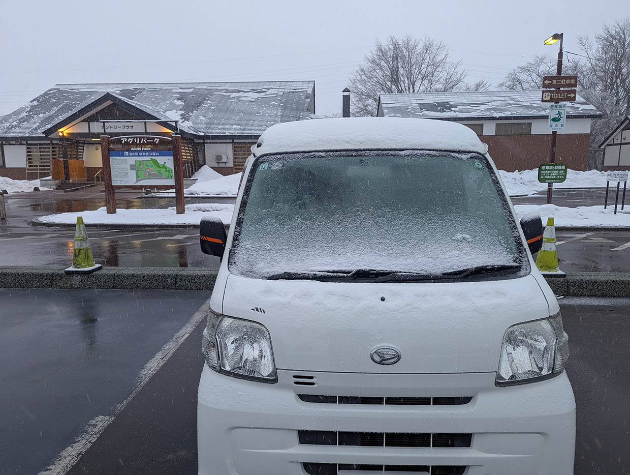 雪が積もった白い軽バンが道の駅なかさつないの駐車場に停まっている