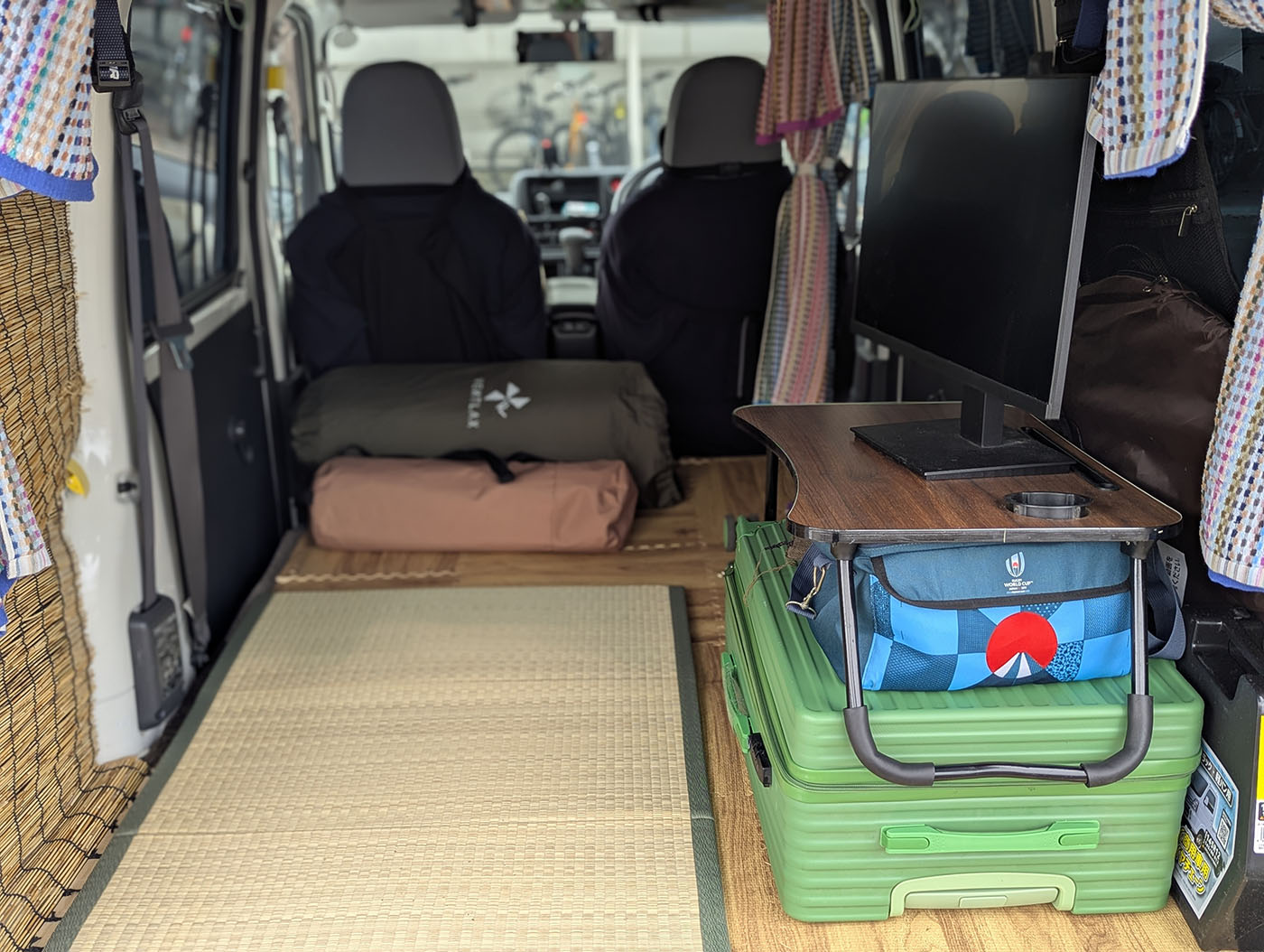 車中泊仕様の軽バン車内に畳マットが敷かれ、寝袋、モニター、テーブル、荷物が整然と配置された快適な旅の準備が整った様子。