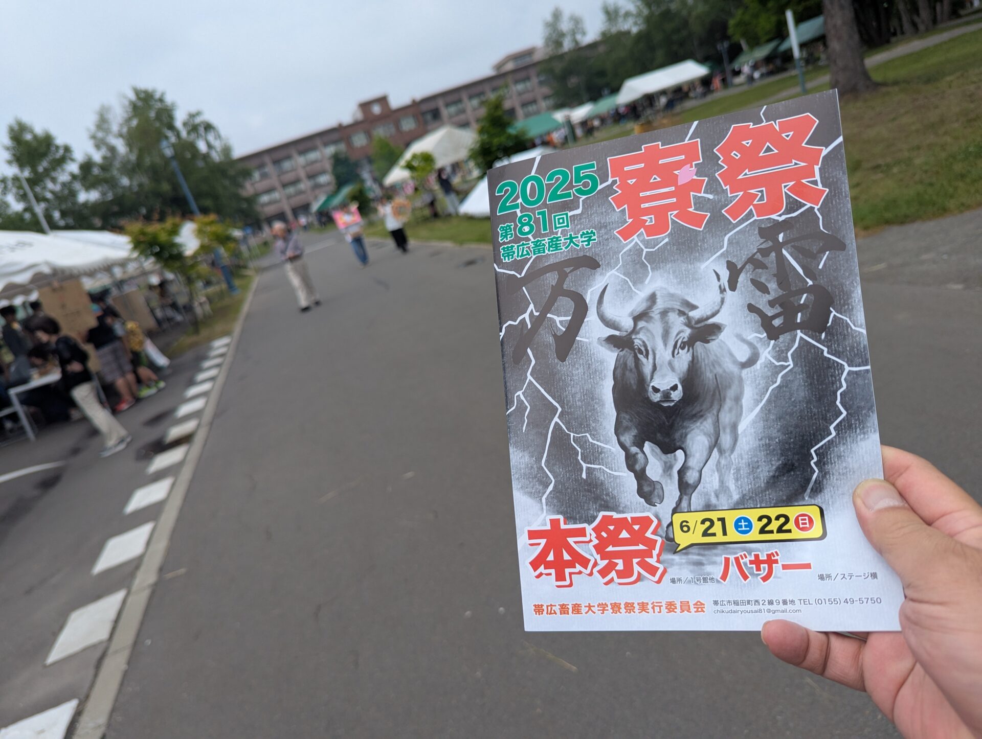 帯広畜産大学のキャンパス内で開催されている第81回寮祭の様子。手前には雷を背景に牛が描かれたインパクトのあるチラシが映り、通路沿いには模擬店のテントや来場者が並ぶ。