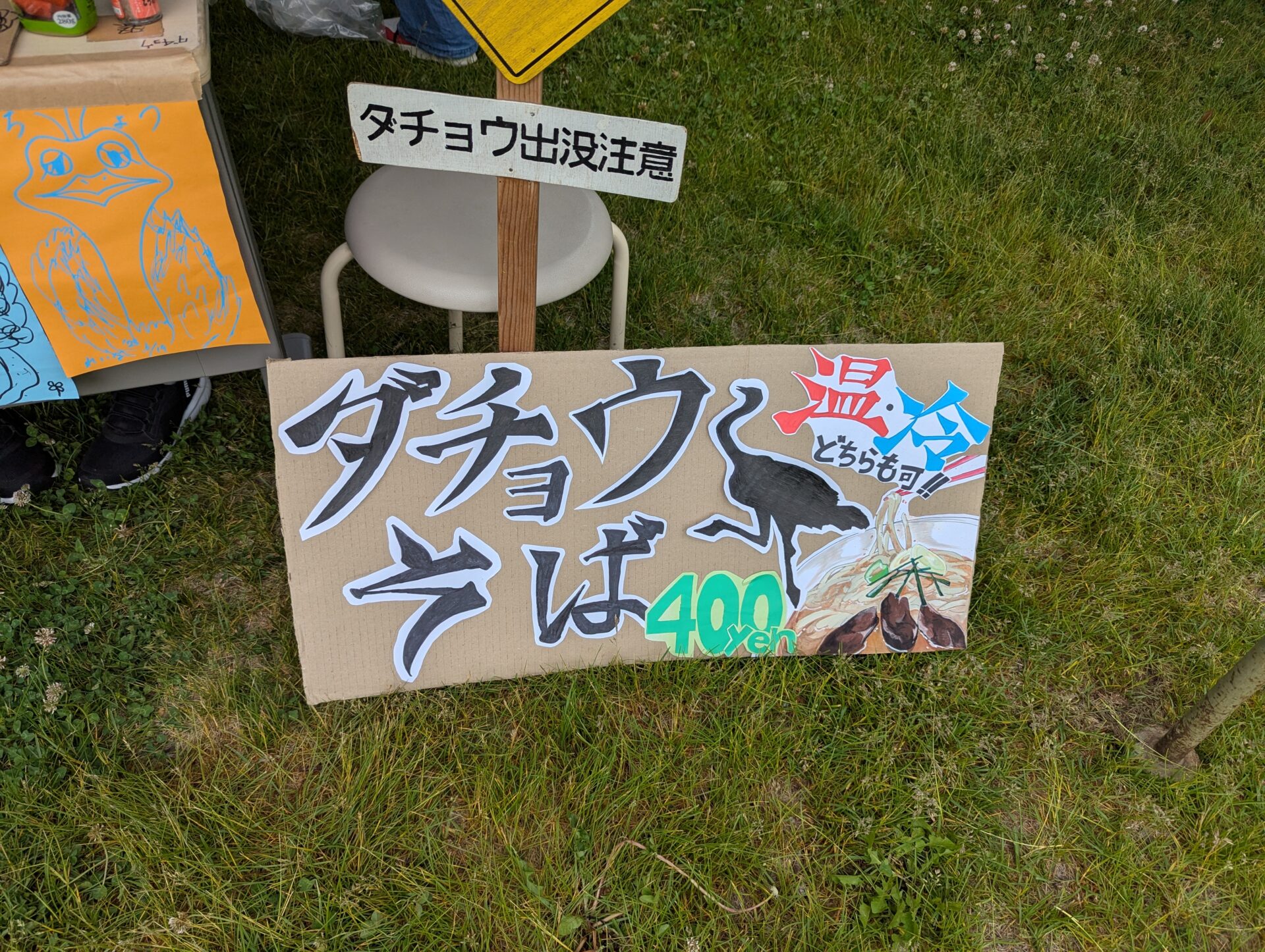 芝生の上に置かれた手作りの『ダチョウそば』の看板。『温・冷どちらも可』と書かれ、400円で販売されている。近くには“ダチョウ出没注意”のユーモラスな標識と手描きのイラストも添えられている。