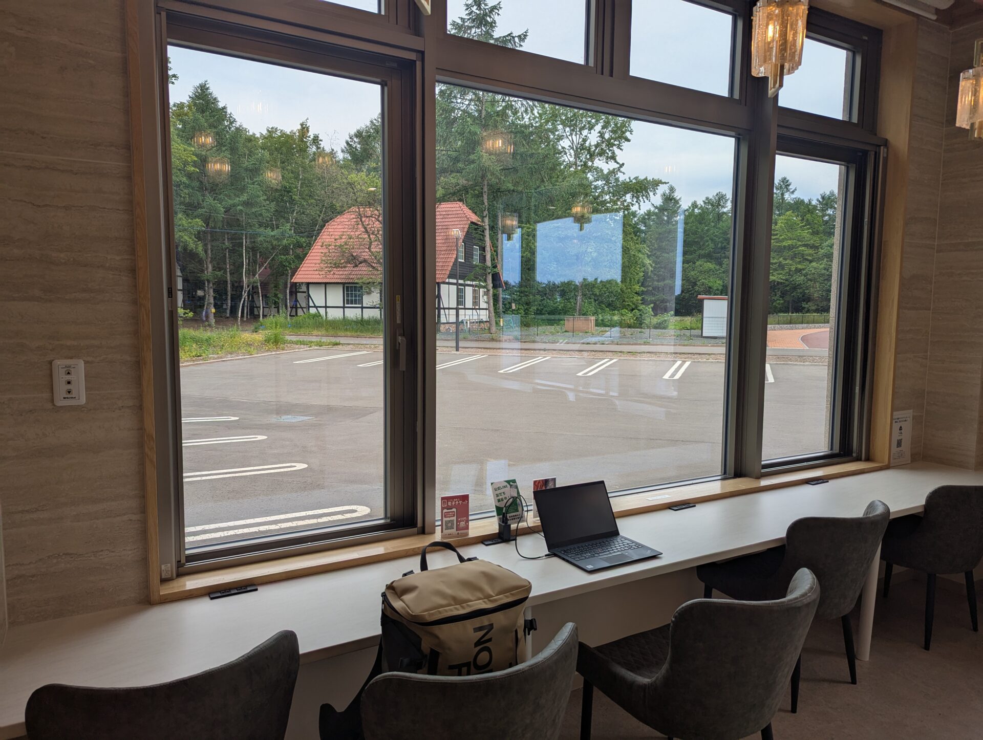 十勝エアポートスパ SORAの休憩スペース。窓際のカウンター席にノートパソコンとバックパックを置き、外の森と赤い屋根の建物が見える。