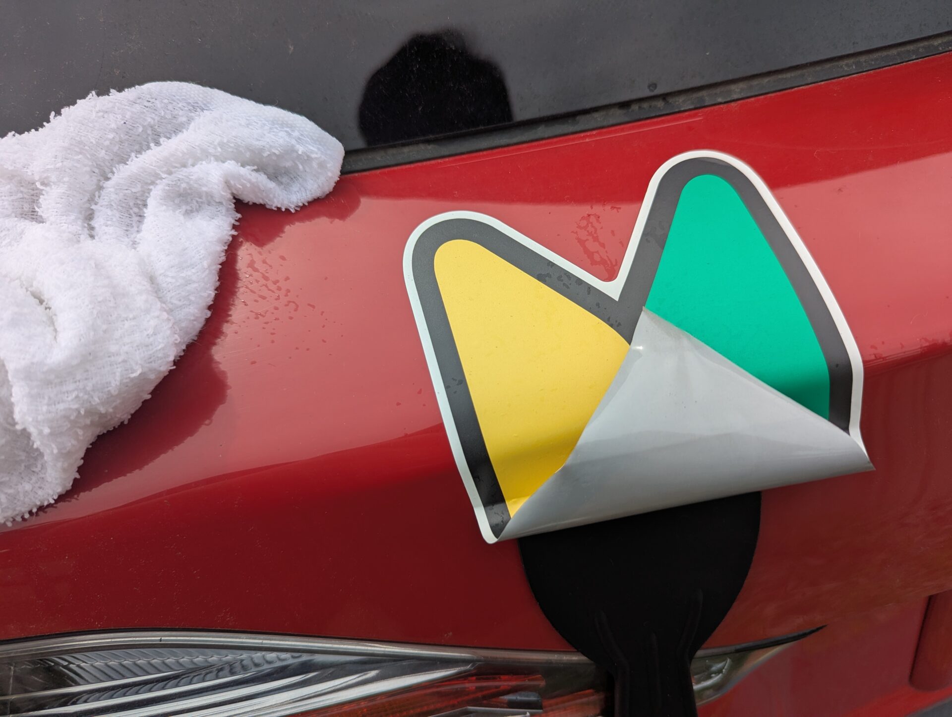 赤い車のリアに貼られた初心者マークを、スクレーパーと白いタオルを使って丁寧にはがしている様子。運転初心者期間の終わりを象徴する一幕。