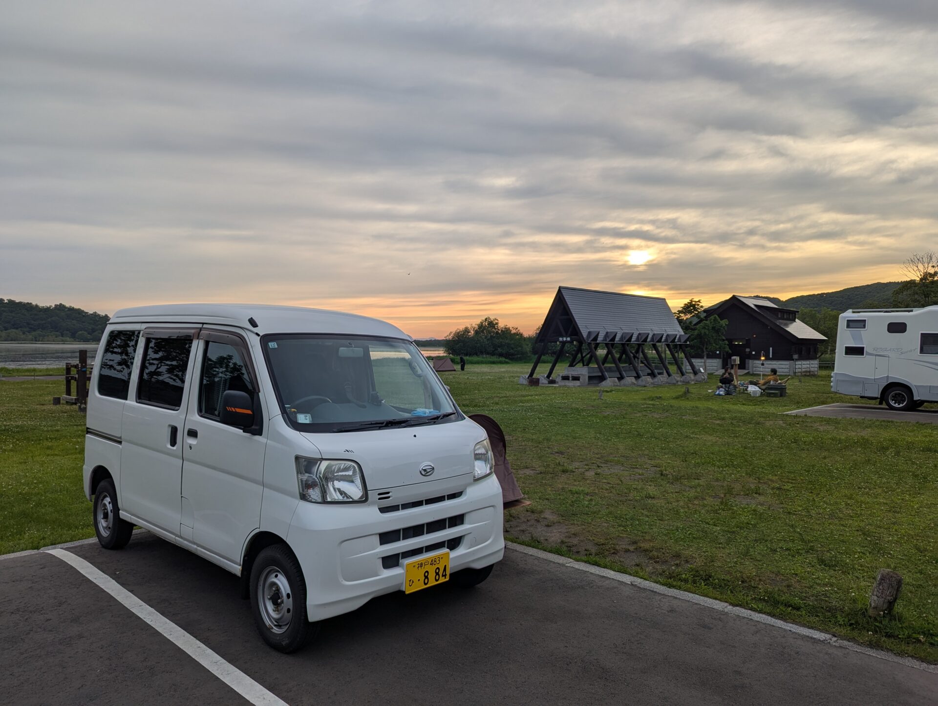 釧路にある達古武キャンプ場の夕方、芝生のサイトに停めた白いハイゼットカーゴと、奥には炊事棟と木造キャビンが並ぶ穏やかな風景。