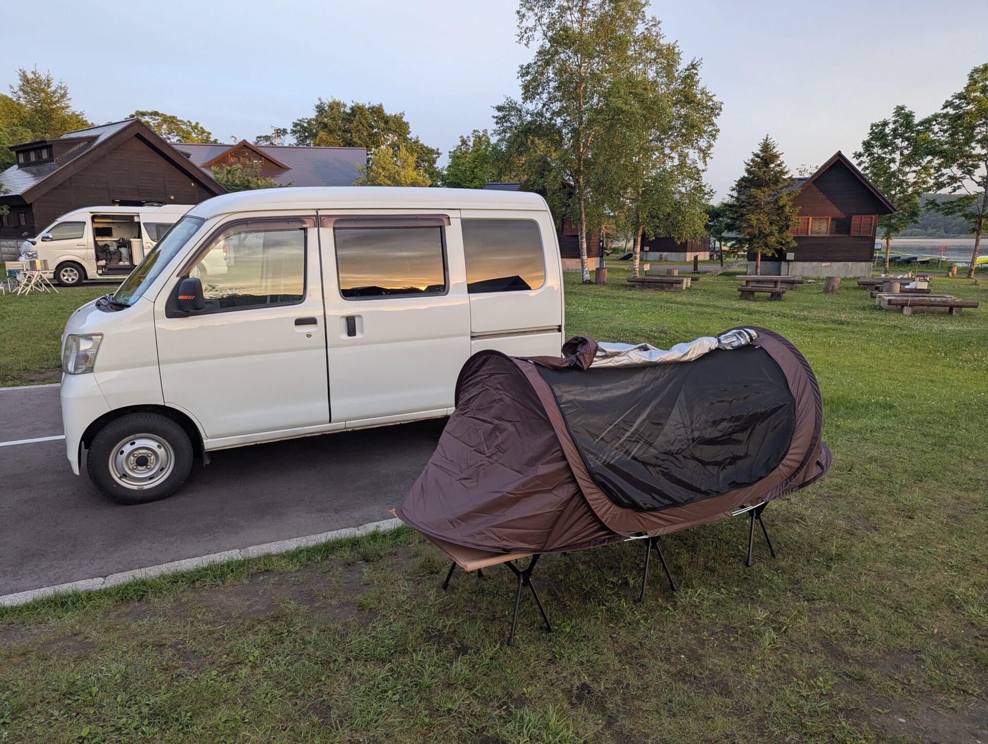 釧路の達古武キャンプ場にて、白いハイゼットカーゴの横に設置されたコット上のポップアップテント。背景には炊事棟やログハウスが並び、夕暮れの柔らかな光が辺りを照らしている。