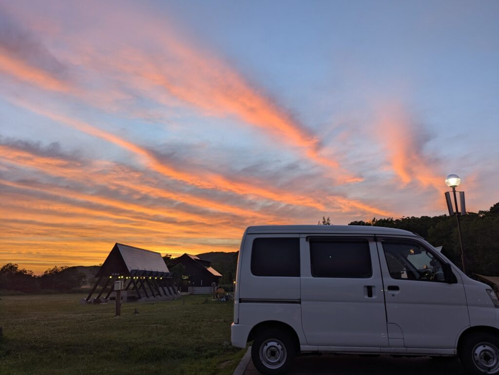 夕焼け空にオレンジの雲が広がる達古武キャンプ場。白いハイゼットカーゴと炊事棟がシルエットになり、ランプが点灯している。