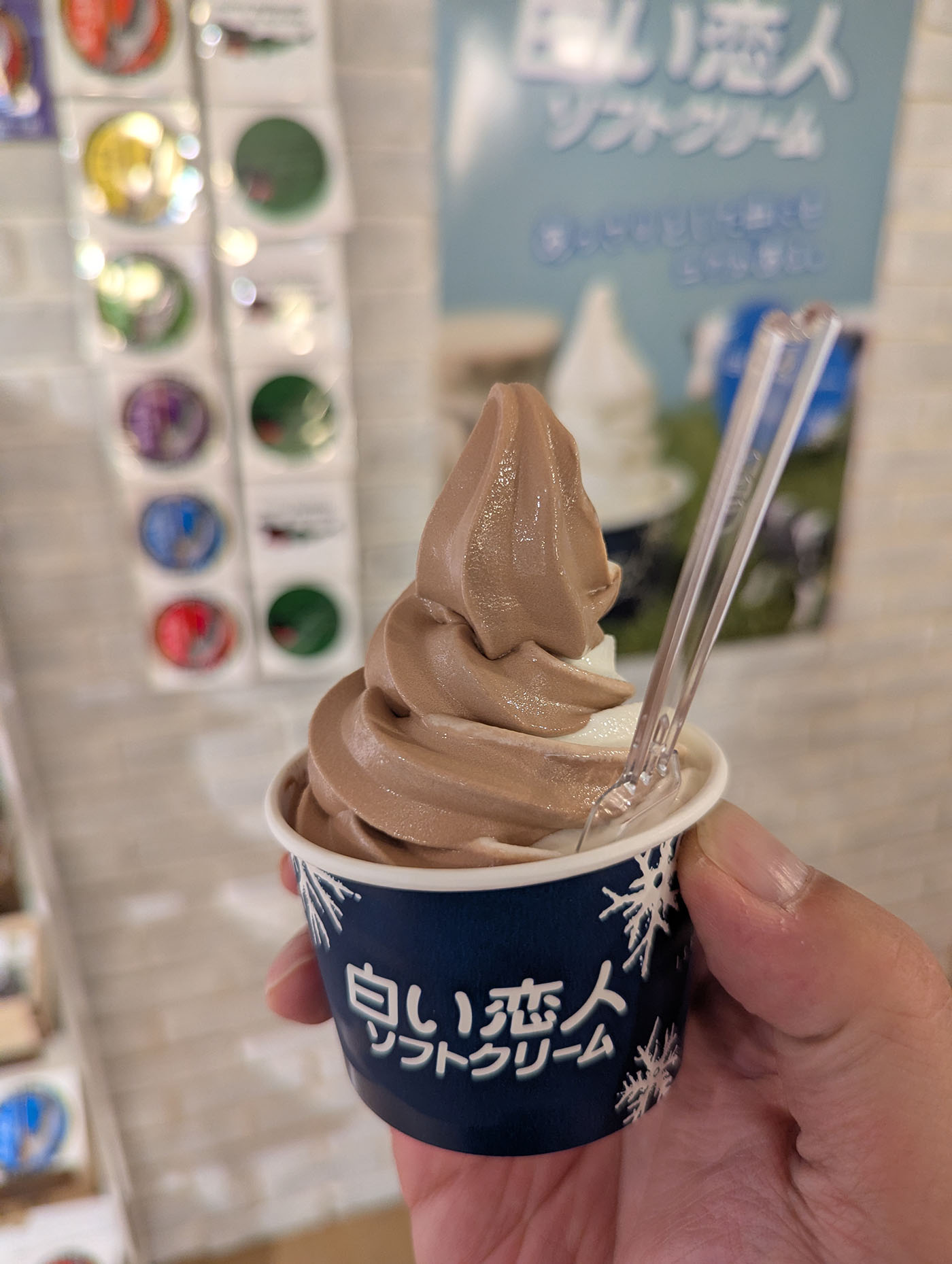 手に持ったカップに盛り付けられた白い恋人のミックスソフトクリーム。チョコとバニラが美しく渦を巻き、背景には「白い恋人ソフトクリーム」と書かれた看板が見える。