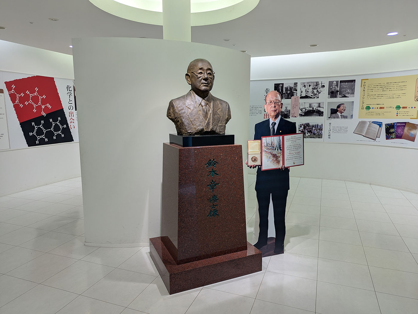 
鈴木章博士の胸像と等身大パネル。ノーベル賞受賞の証書やメダルを持ったパネルが胸像の隣に立ち、背景には化学の功績を紹介するパネル展示が並ぶ。