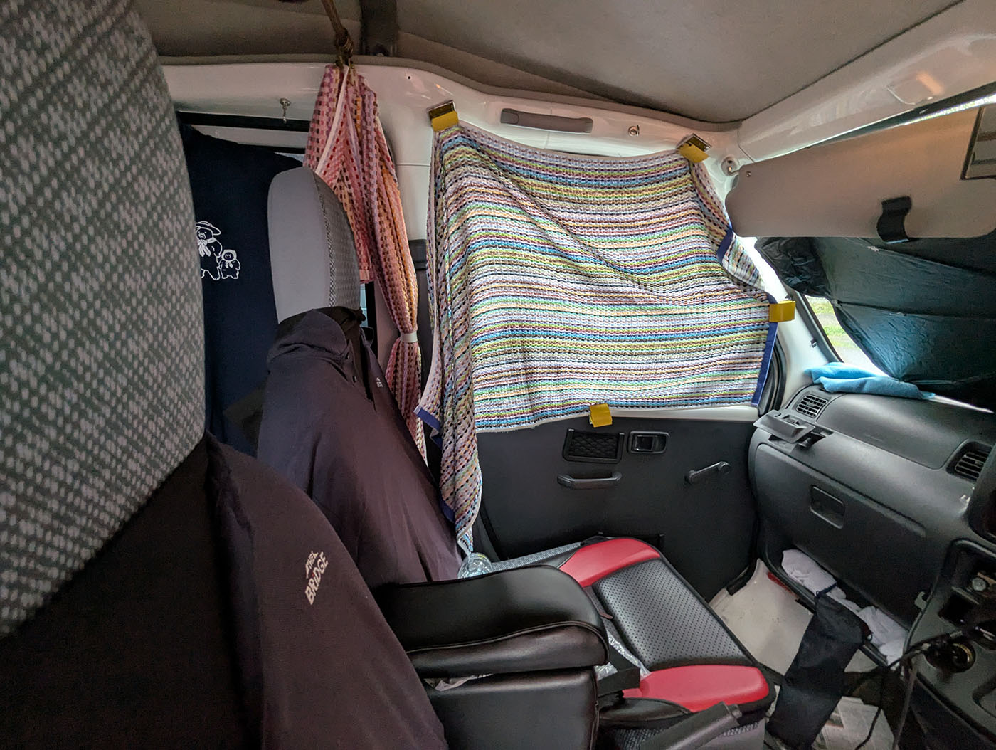 車中泊仕様の車内を前方から撮影した写真。運転席と助手席の窓にはカラフルなブランケットやカーテンが貼られ、日差しや視線を遮るように工夫されている。暗く落ち着いた空間が確保され、長時間駐車や仮眠に備えた遮光対策がしっかりと施されている様子。