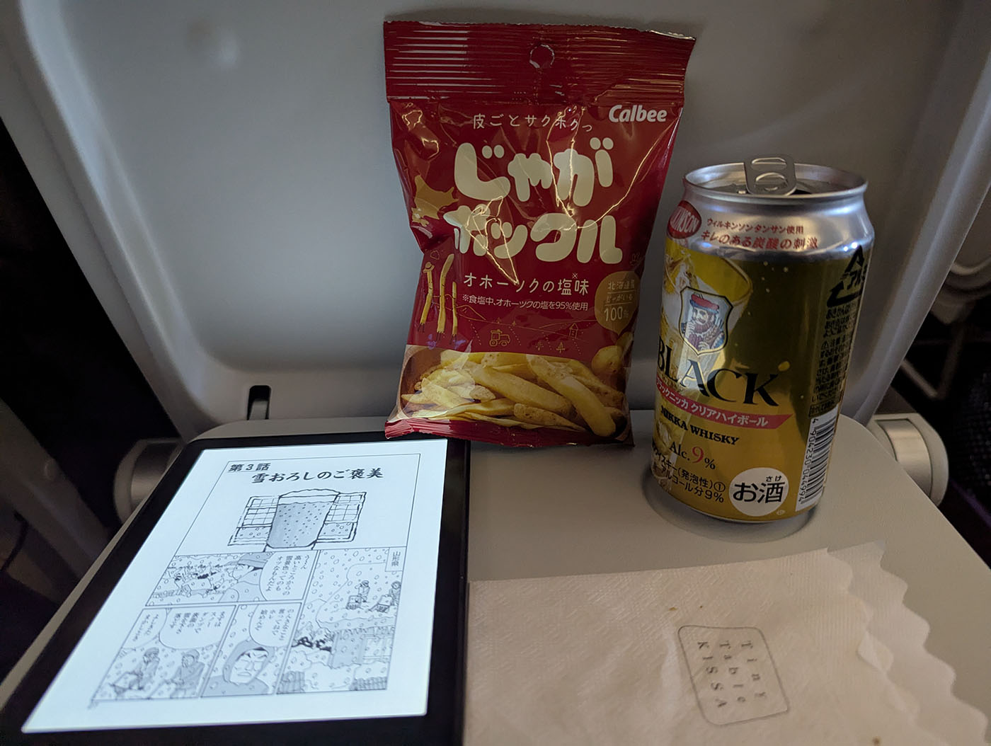 飛行機のテーブルに並べられたじゃがポックルの袋と缶のハイボール、そして電子書籍リーダー。20分の遅れの間に、北海道の名物おつまみとお酒を楽しみながら、帰りの機内でほっとひと息つく様子が伝わる。