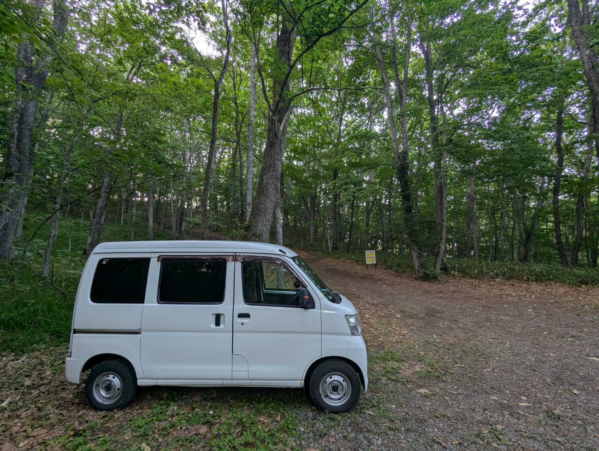 一本木展望タワーの駐車場として使われている山のスペースに停めた白い軽バン。周囲は緑に包まれ、未舗装の地面に落ち葉が散らばる。