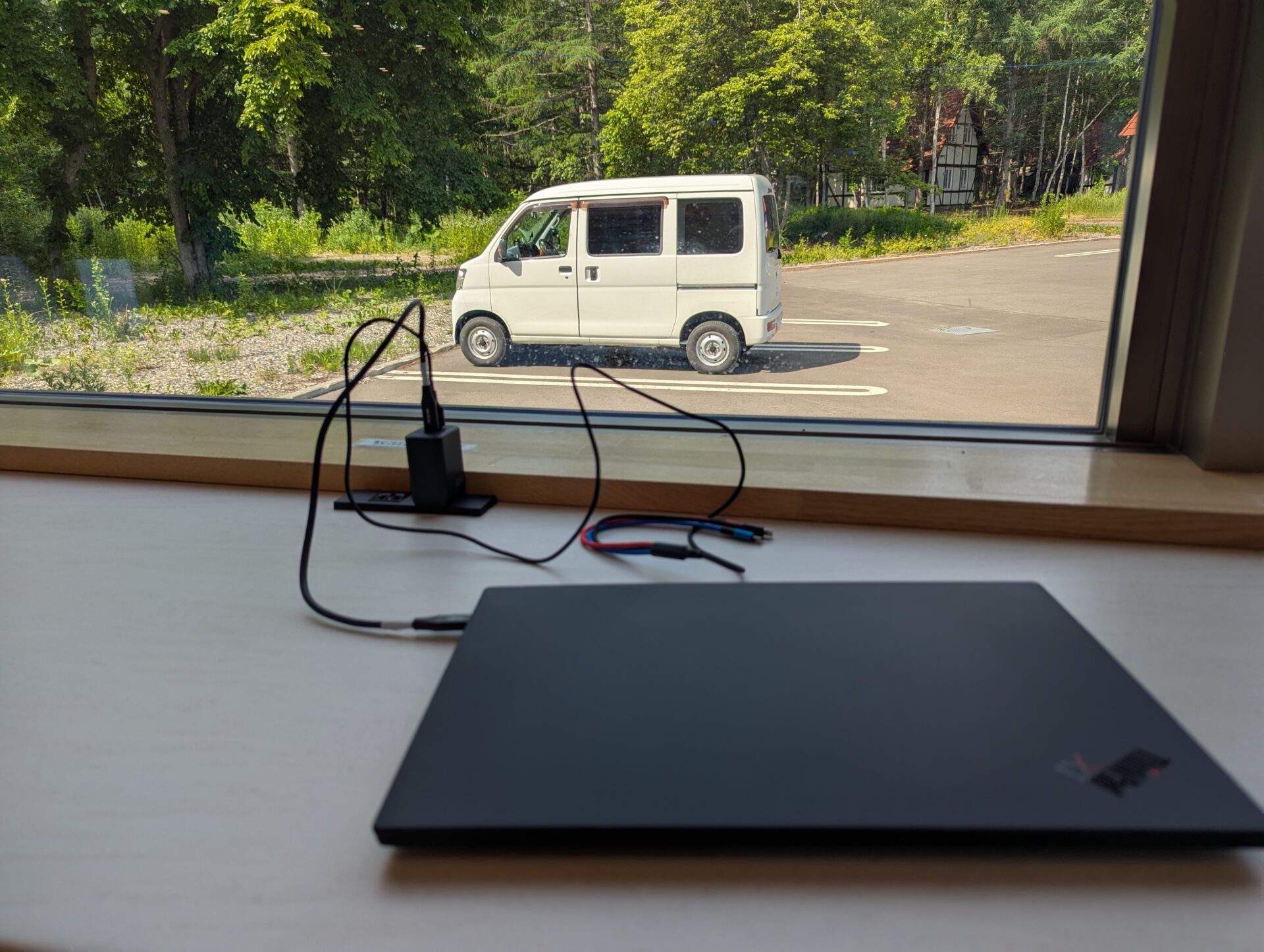 十勝エアポートスパそらの休憩所のカウンター席にノートパソコンを置き、窓の外に停めた白い軽バンを眺めながら作業する風景。木々に囲まれた駐車場と静かな空間が広がる。