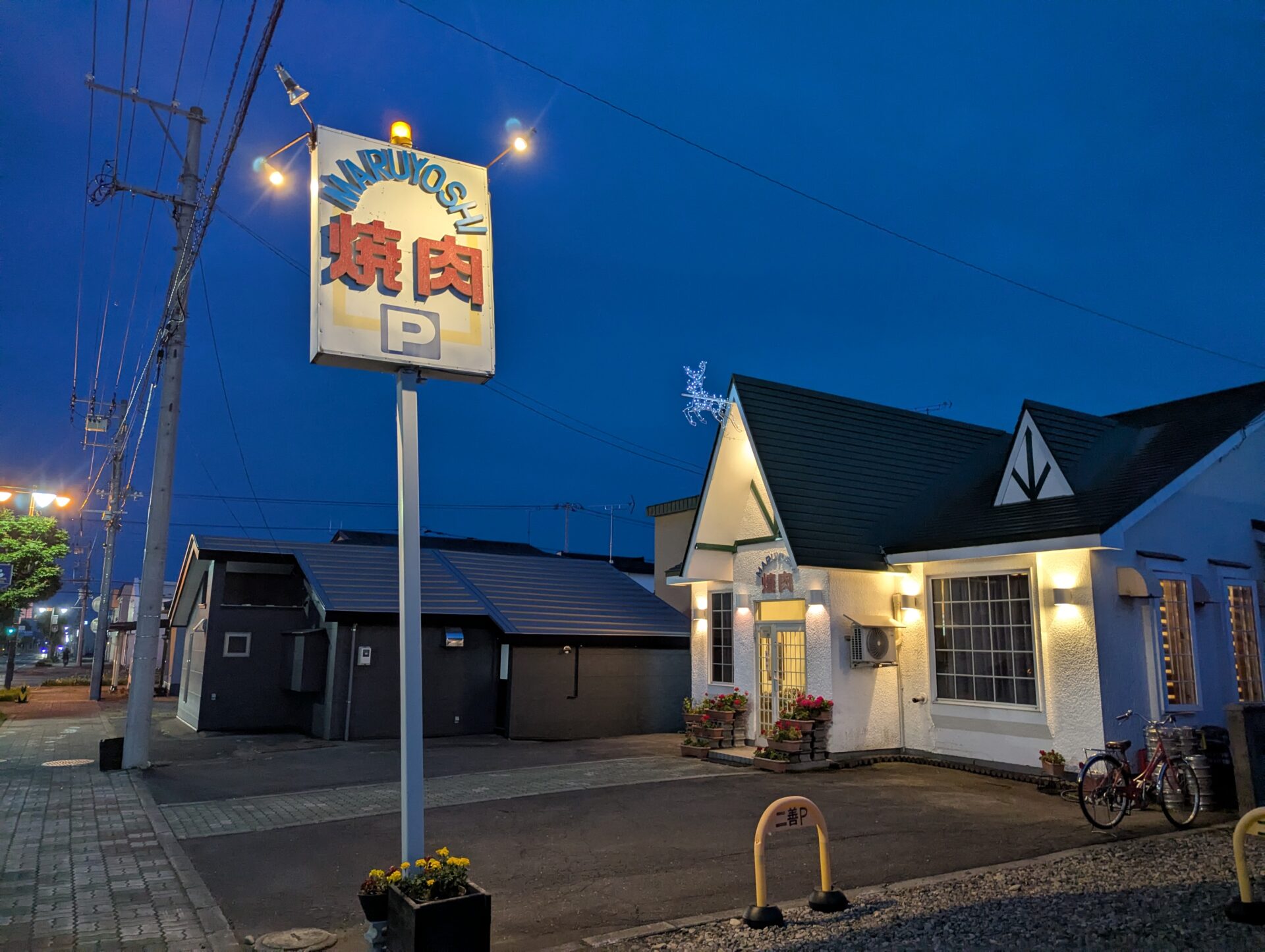 夜の鹿追の街角に建つ「焼肉まるよし」の外観。白い壁と三角屋根が特徴で、青い夜空の下、看板と建物が温かい光に照らされている。