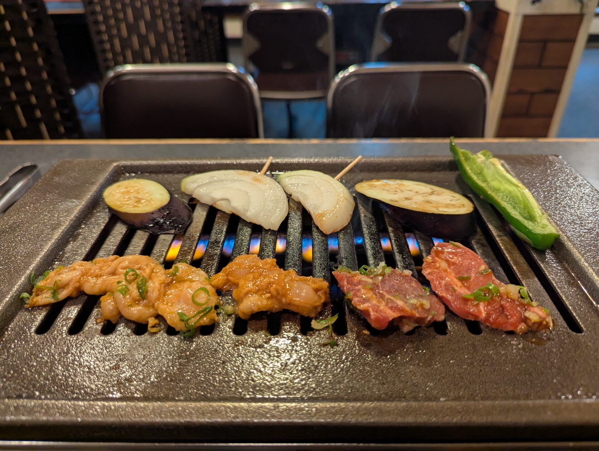 焼肉まるよしのテーブルで、茄子や玉ねぎ、ピーマン、味付けされた肉が網の上で焼かれている様子。炭火の香りが伝わってくるよう。