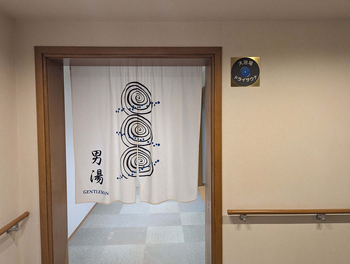 新冠温泉の男湯入り口。白い暖簾に渦巻き模様が描かれ、右上には『大浴場 ドライサウナ』の案内板がある
