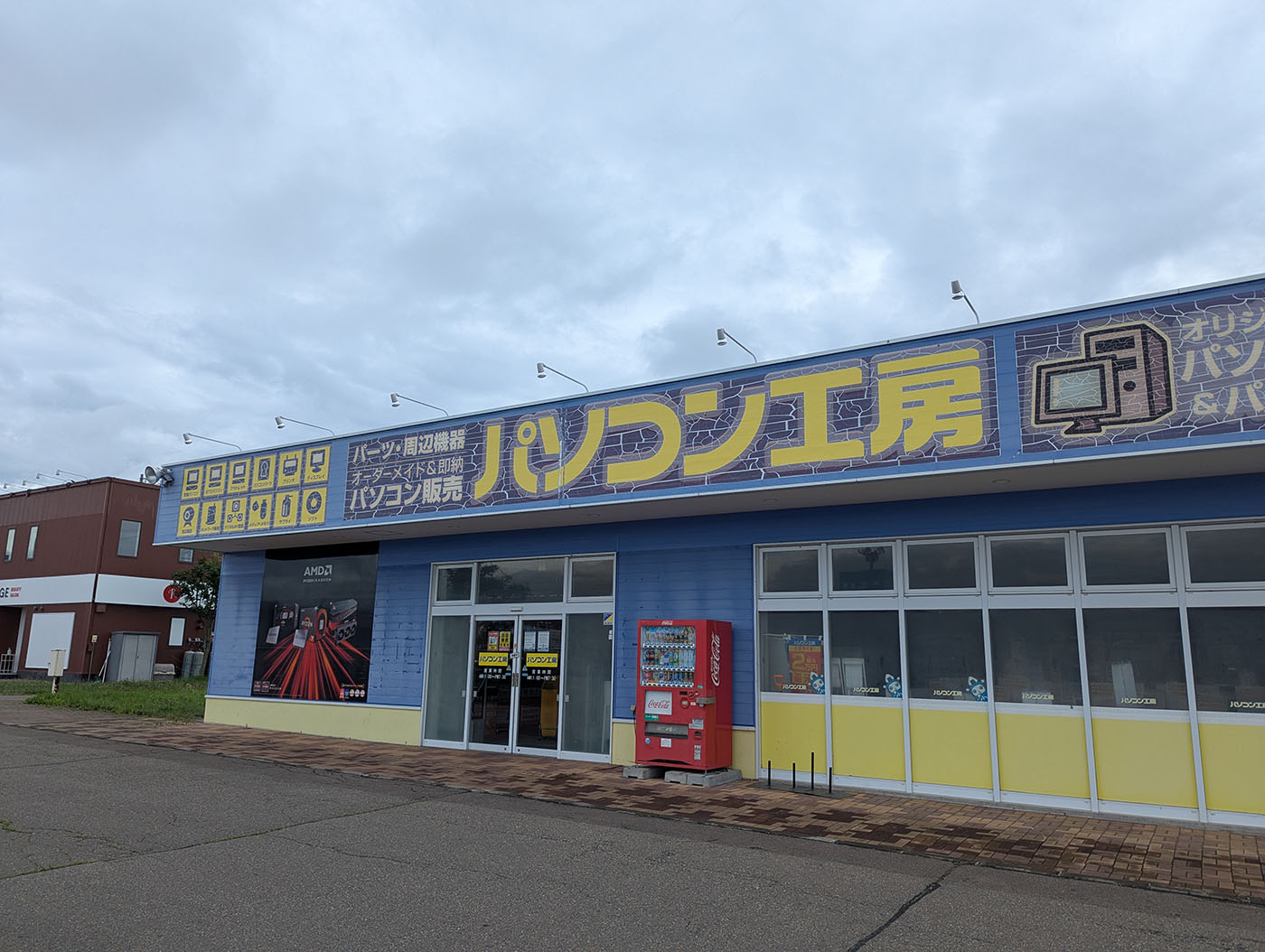 帯広の「パソコン工房」の店舗外観。黄色と青の外壁が特徴で、自動販売機も設置されている。