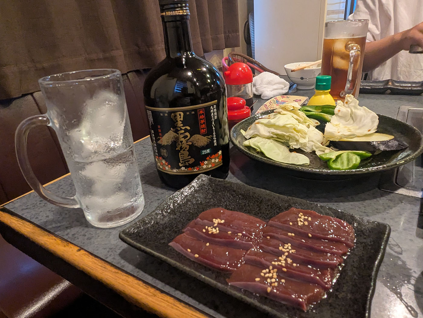 焼肉まるよし店内のテーブル。黒霧島のボトルと氷たっぷりのグラス、キャベツやナスの盛り合わせ、そしてレバーが並んでいる。