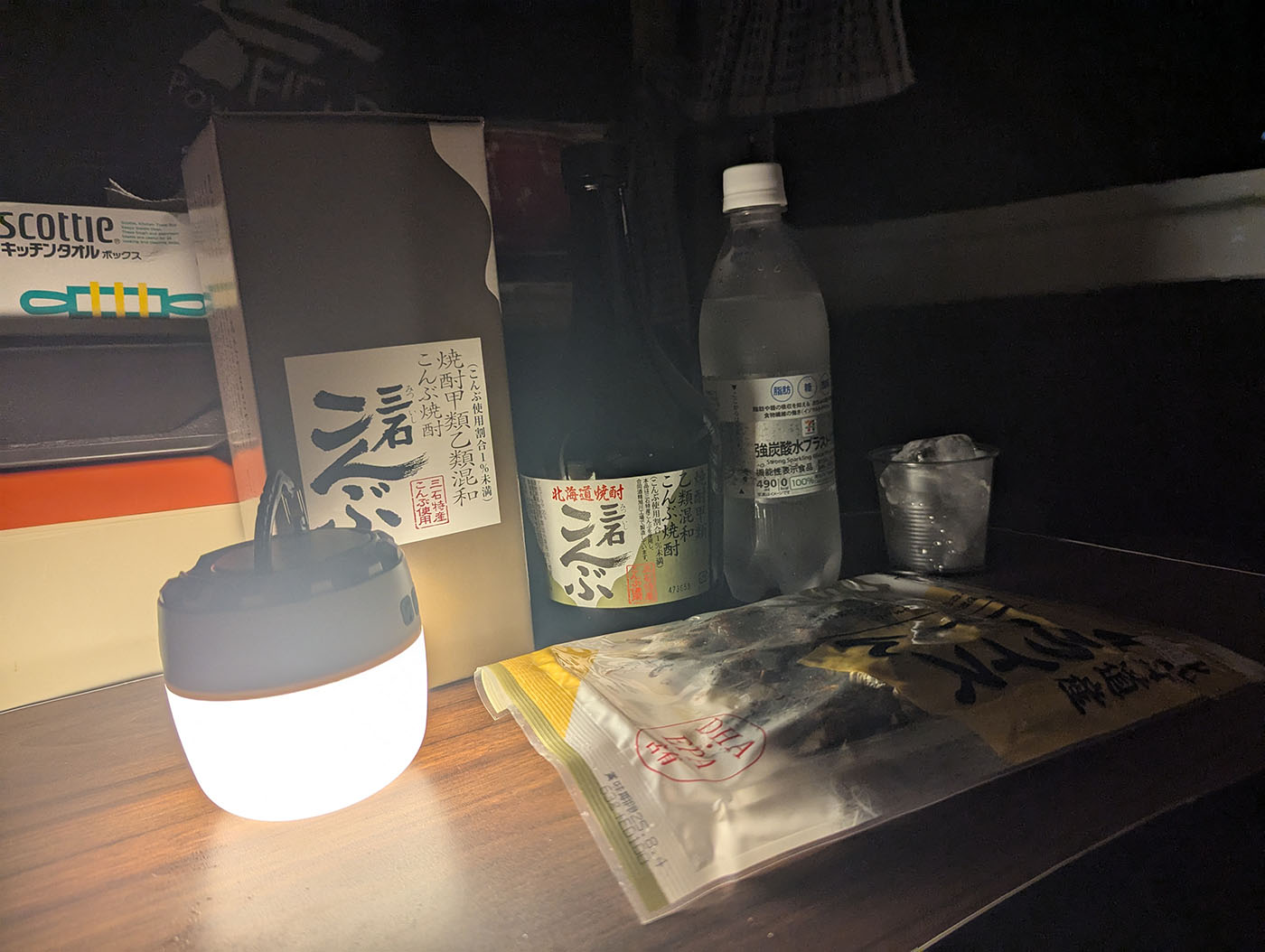 車中泊の車内で、ランタンの灯りに照らされた三石こんぶ焼酎、炭酸水、氷、乾きもののおつまみが並ぶ夜のテーブル。