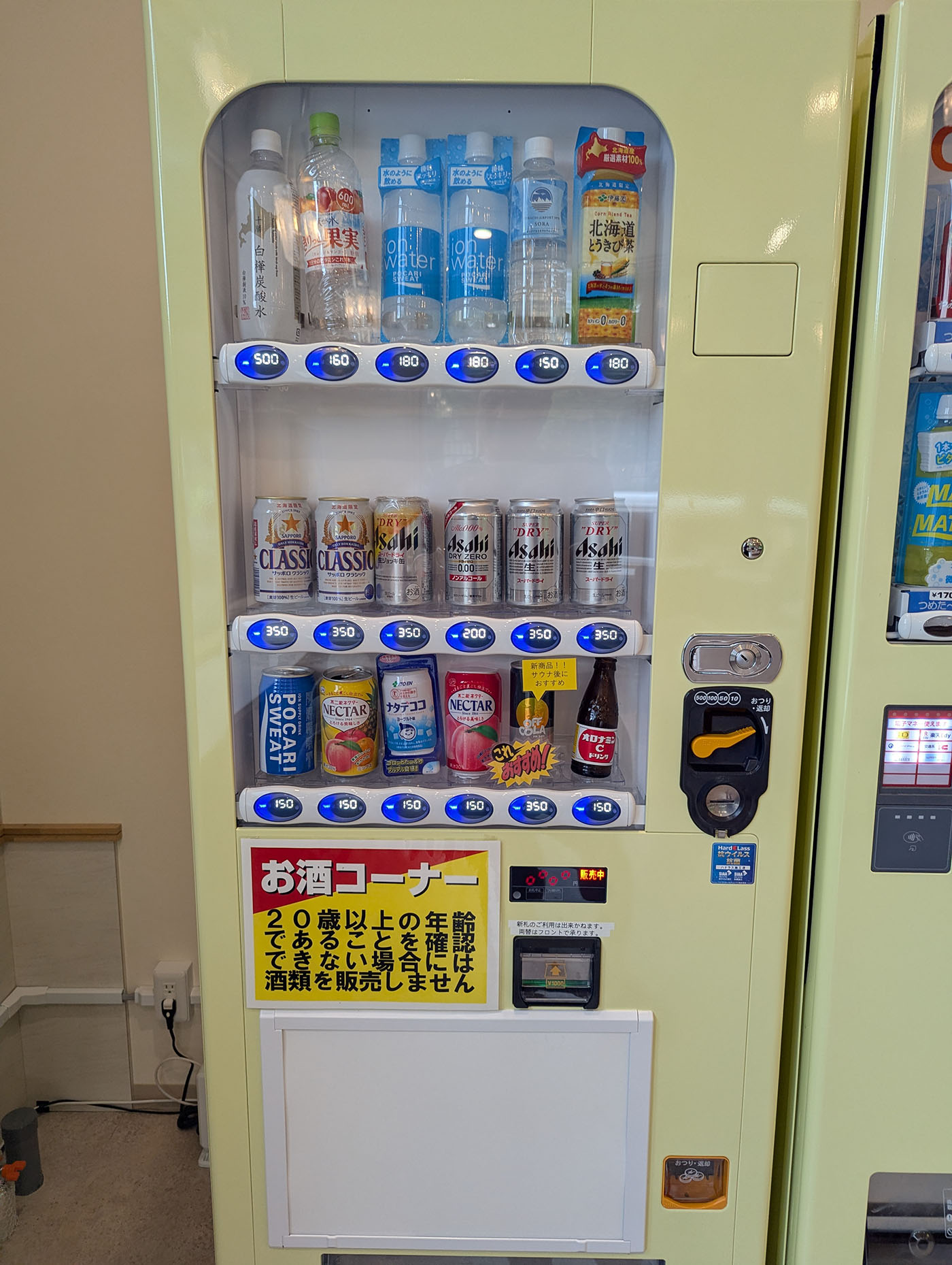 自動販売機に並ぶ水、ジュース、ビールなどの飲料。20歳以上確認の「お酒コーナー」表示も見える。