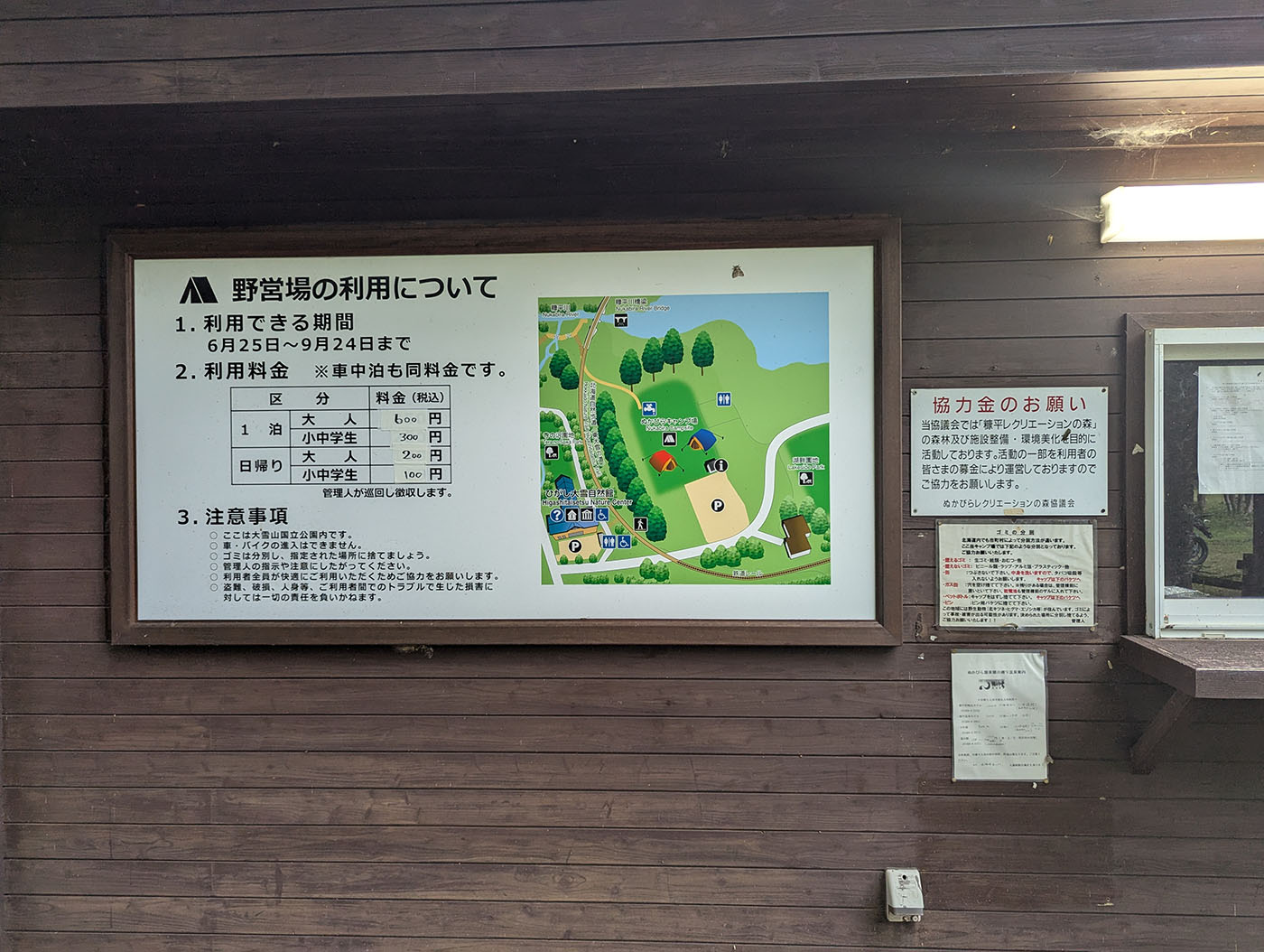糠平野営キャンプ場の利用案内板と料金表、サイトマップが掲示された管理棟の壁面