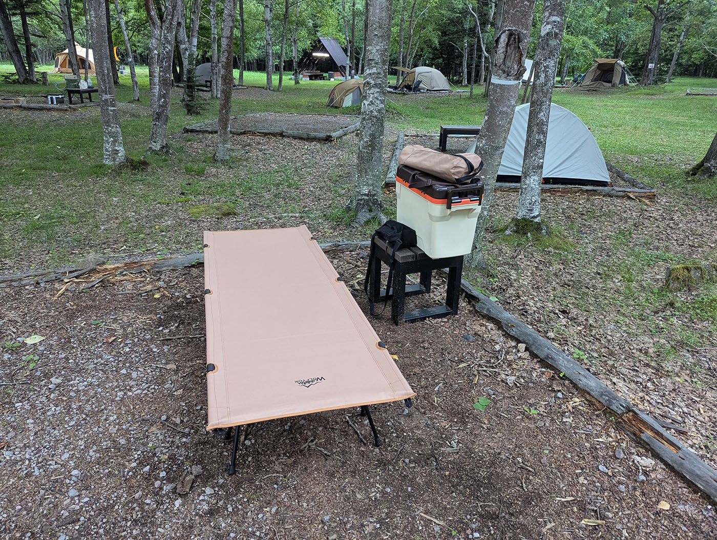 林間のキャンプサイトに、ワンタッチコットと収納コンテナ、サイドテーブルのみが設置された状態。周囲には他のテントも点在している。