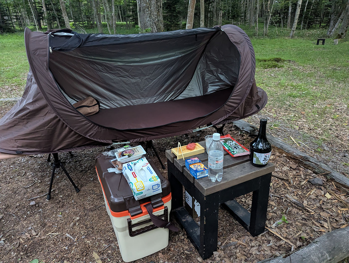 林間のキャンプ場に設置された茶色のソロ用テントとコット。手前のテーブルには北海道焼酎「こんぶ」と氷入りグラス、たらこやチーズのおつまみ、炭酸水、ティッシュや食品類が並べられている。