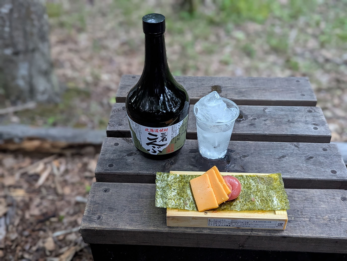 木のテーブルの上に置かれた北海道産焼酎「二海 こんぶ」、ロックグラス、チーズとたらこを海苔にのせたおつまみ。