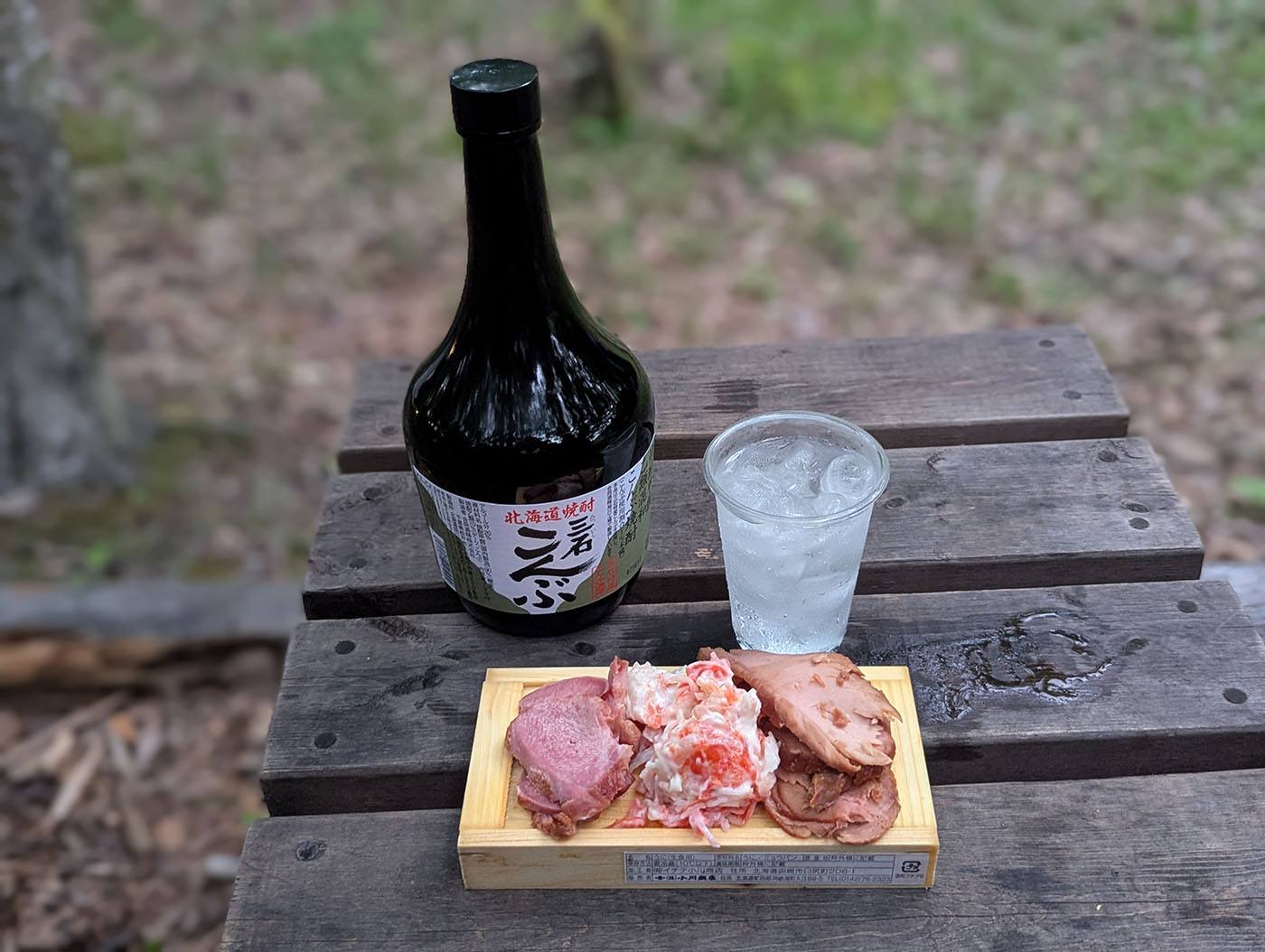 木のテーブルの上に並んだ北海道焼酎「三石 こんぶ」、氷入りのグラス、タン、かにかま、チャーシューのおつまみセット。