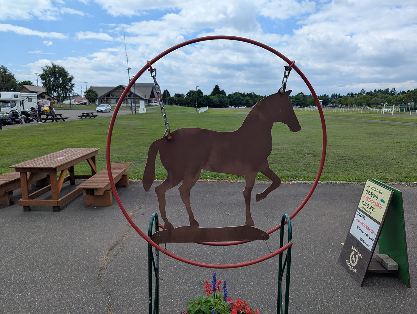 赤い円形フレームに吊るされた馬のシルエット看板。背景には芝生広場とベンチ、遠くに建物や駐車場が見える。