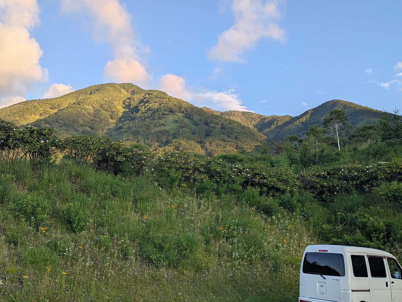 ヌプカの里で迎えた朝。朝日が山の斜面をやさしく照らし、深い緑がしっとりと浮かび上がる。右手には白い軽バンが停まり、静かな車中泊の朝を物語っている。