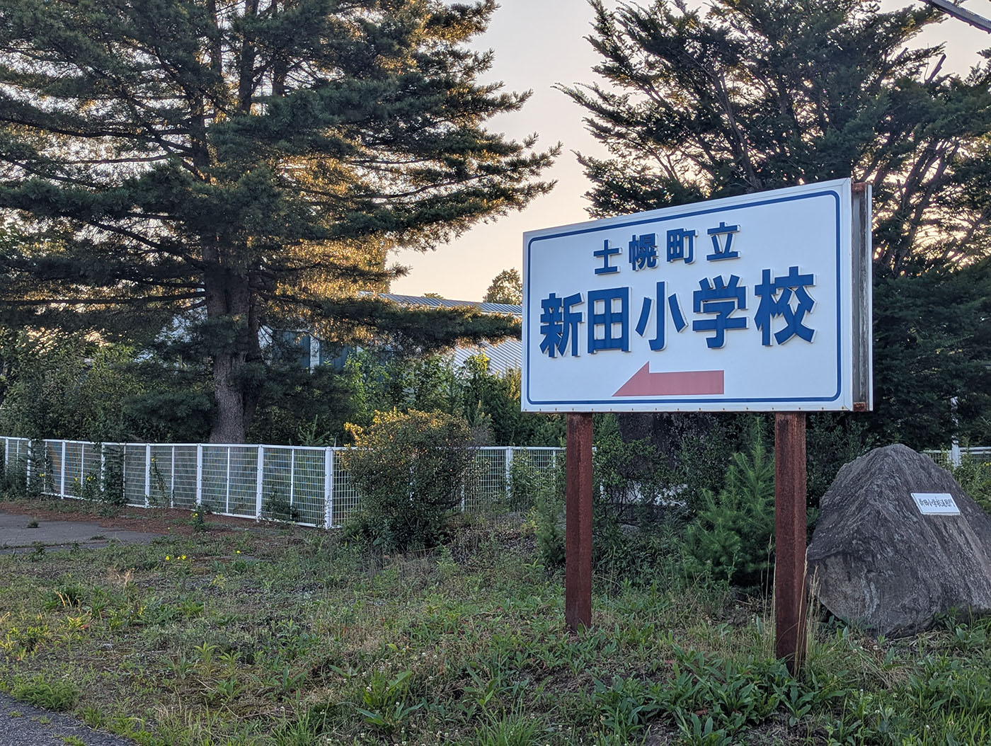 北海道・士幌町にある「新田小学校」の看板。白地に青い文字で校名が書かれ、赤い矢印が進行方向を示している。背後には大きな針葉樹が並び、夕方の光が差し込んでいる。