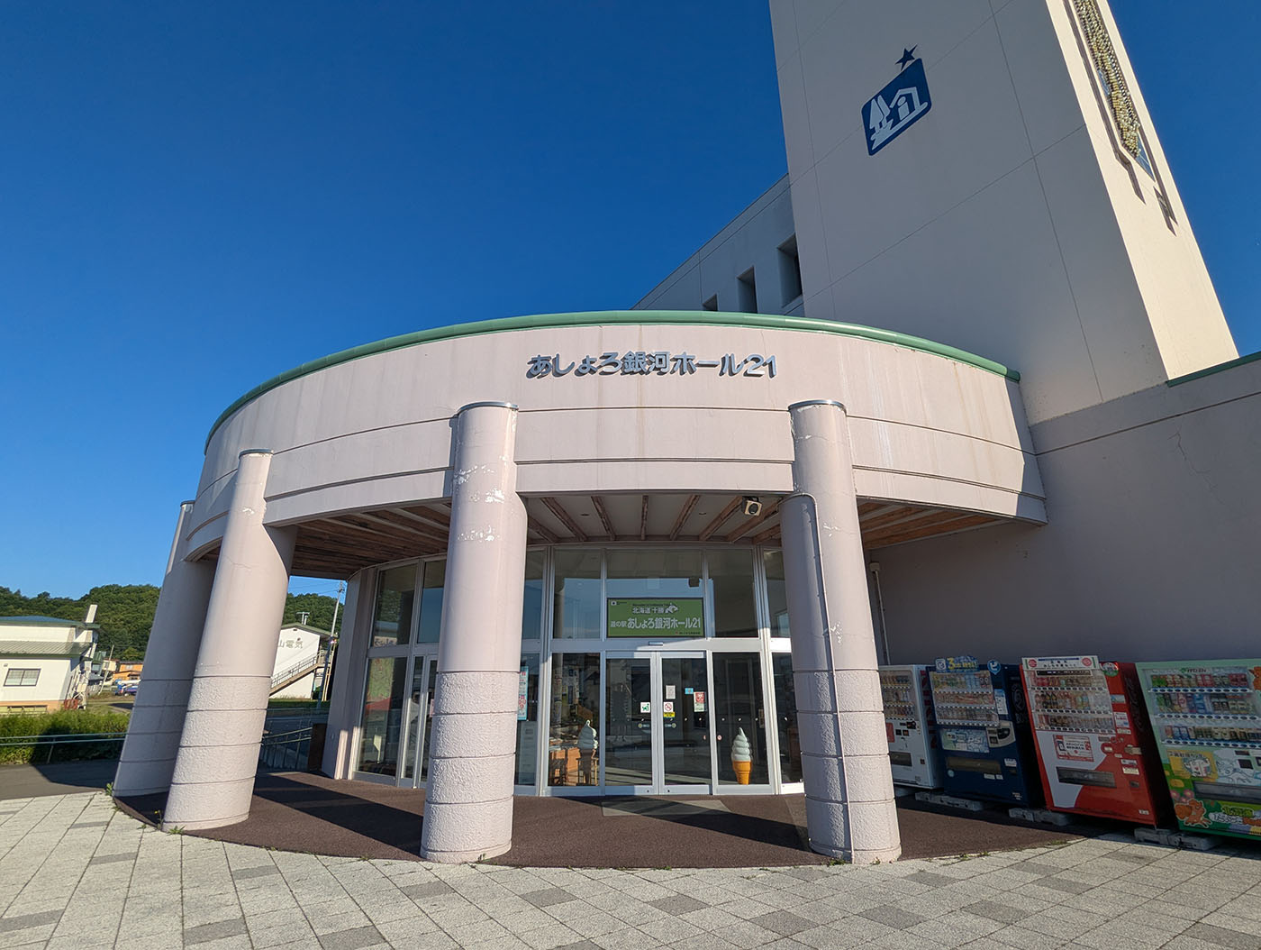 北海道足寄町にある「道の駅あしょろ 銘酒ホール21」の外観。円形のエントランスと白い外壁が特徴的で、自動販売機が並び、青空の下に立つ堂々とした建物。