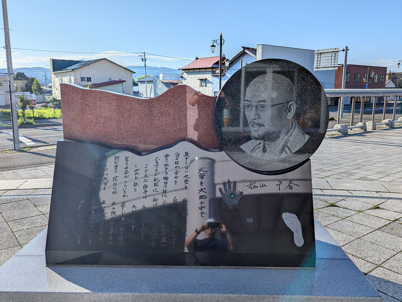 北海道足寄町にある松山千春の記念碑。石碑には松山千春の顔が刻まれた円形のレリーフとともに、手形・足形・直筆サインが彫られている。詩が刻まれたモニュメント越しに、街並みと青空が広がる。