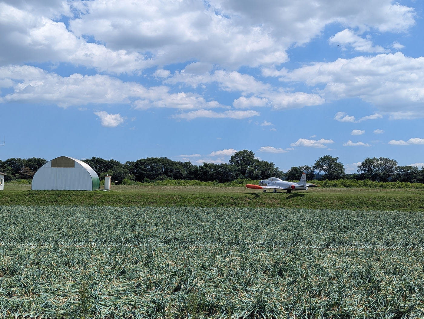 畑の奥に格納庫と並んで置かれたT-33A退役練習機、青空と雲が広がる美幌の風景