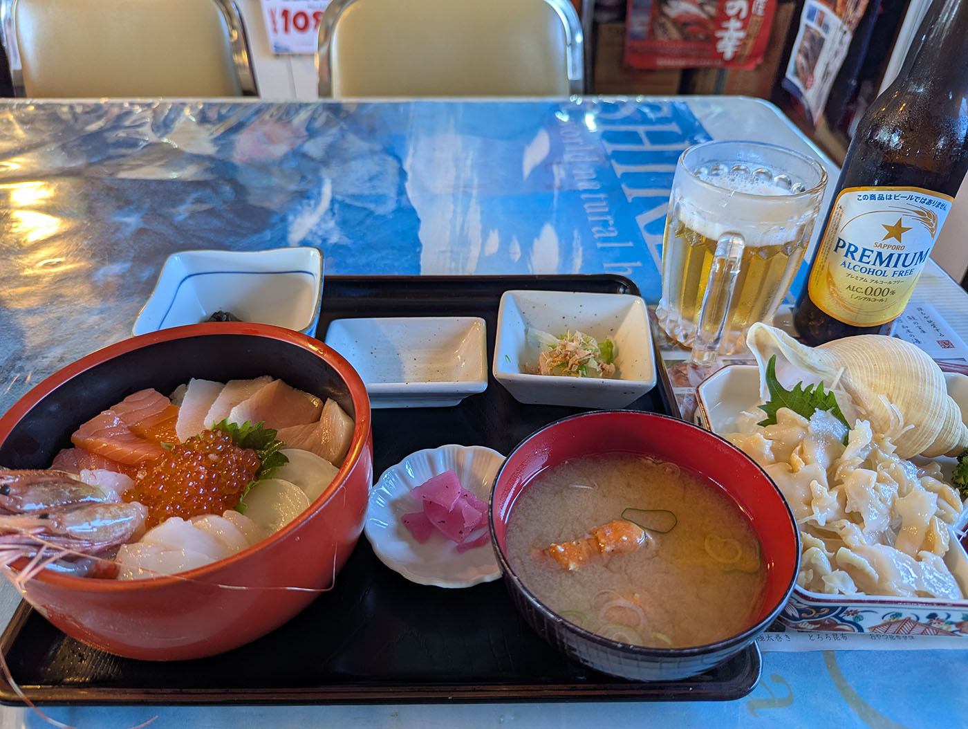 北の国から純の番屋の海鮮丼セット。エビ、サーモン、ホタテ、イクラなどが盛られた丼に、つぶ貝の刺身、小鉢、味噌汁、ノンアルコールビールが並ぶ。