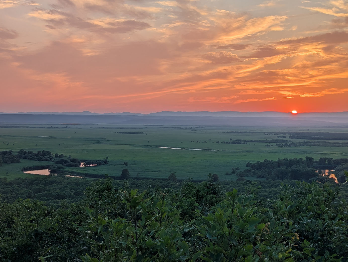 細岡展望台から見た釧路湿原の夕焼け。地平線に沈む太陽と、湿原に流れる川が夕日を反射して輝く広角の風景。