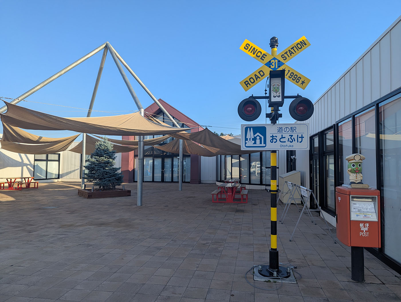 道の駅おとふけの中庭にある踏切風のモニュメントと郵便ポスト。晴天の下、三角形の屋根と日除けが特徴的なモダンな建物が並ぶ。