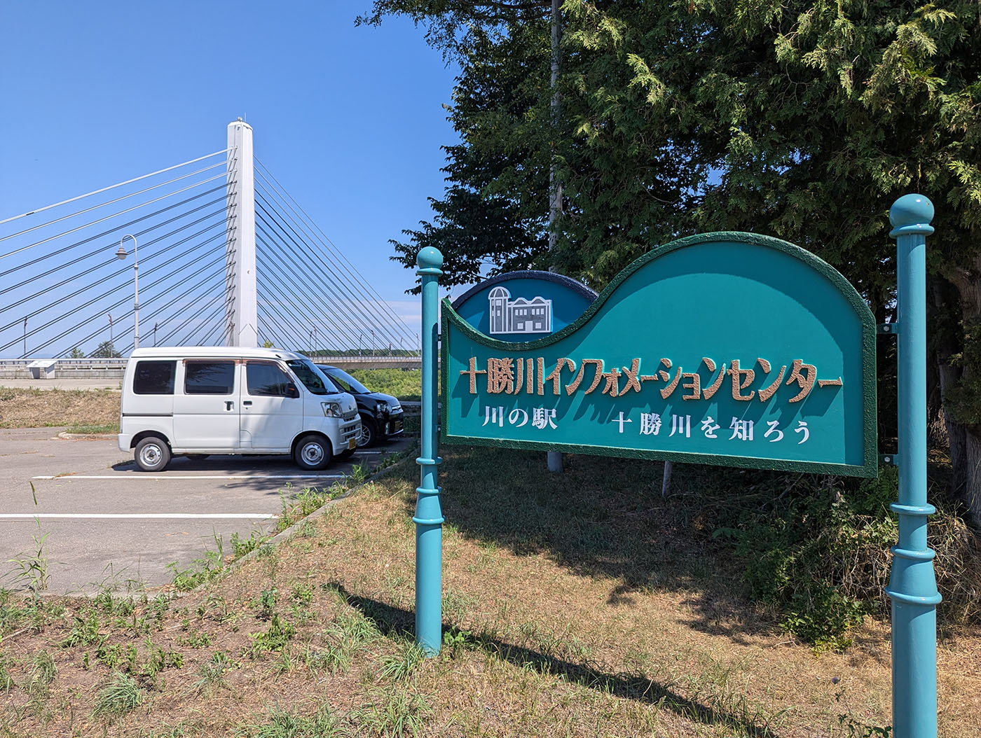 十勝川インフォメーションセンターの看板と、背景に見える白い吊り橋と停車中の軽バン。