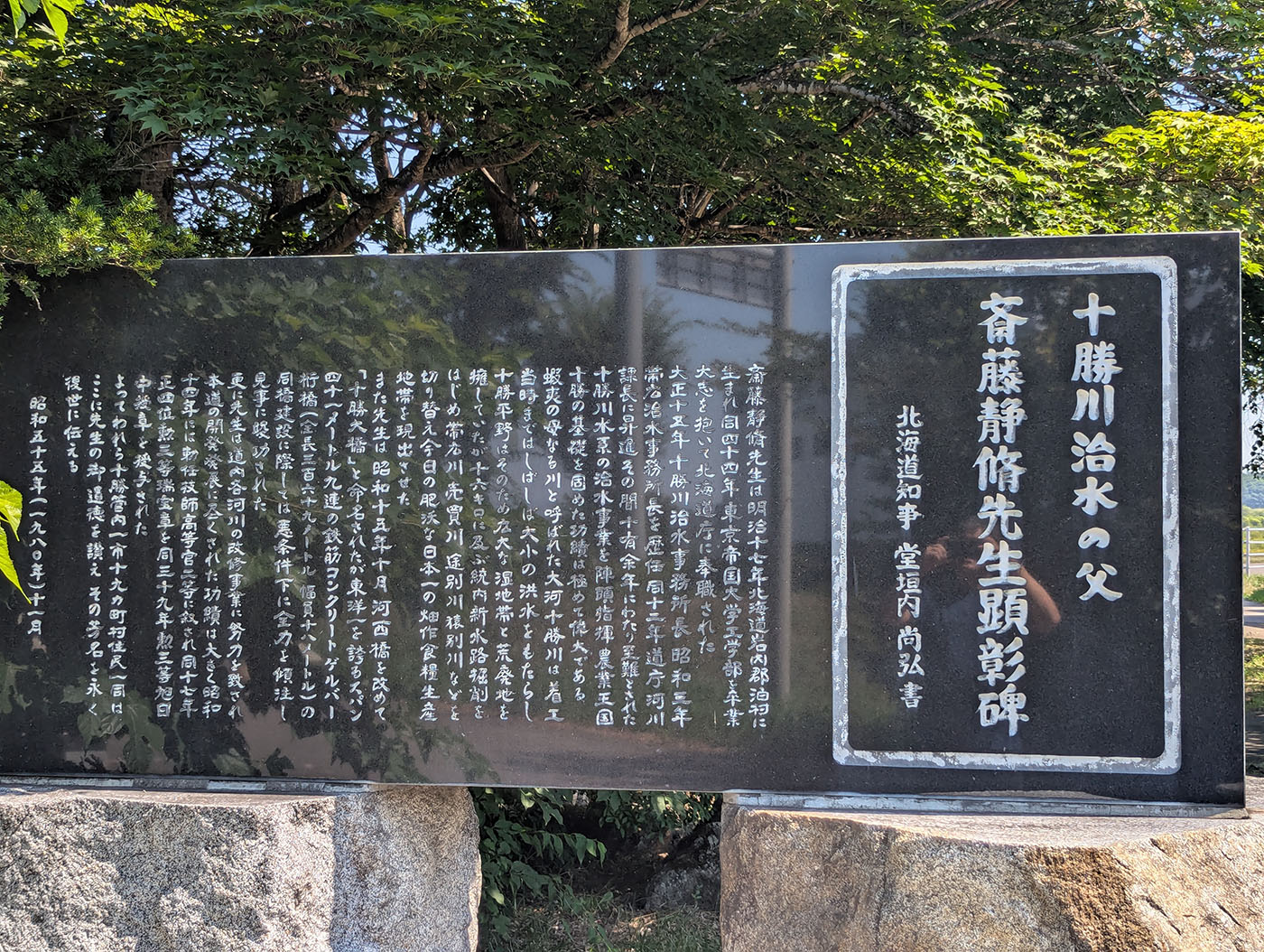 「十勝川治水の父」と称される斎藤静修氏の功績をたたえる黒御影石の顕彰碑。背景には緑の木々が広がる。