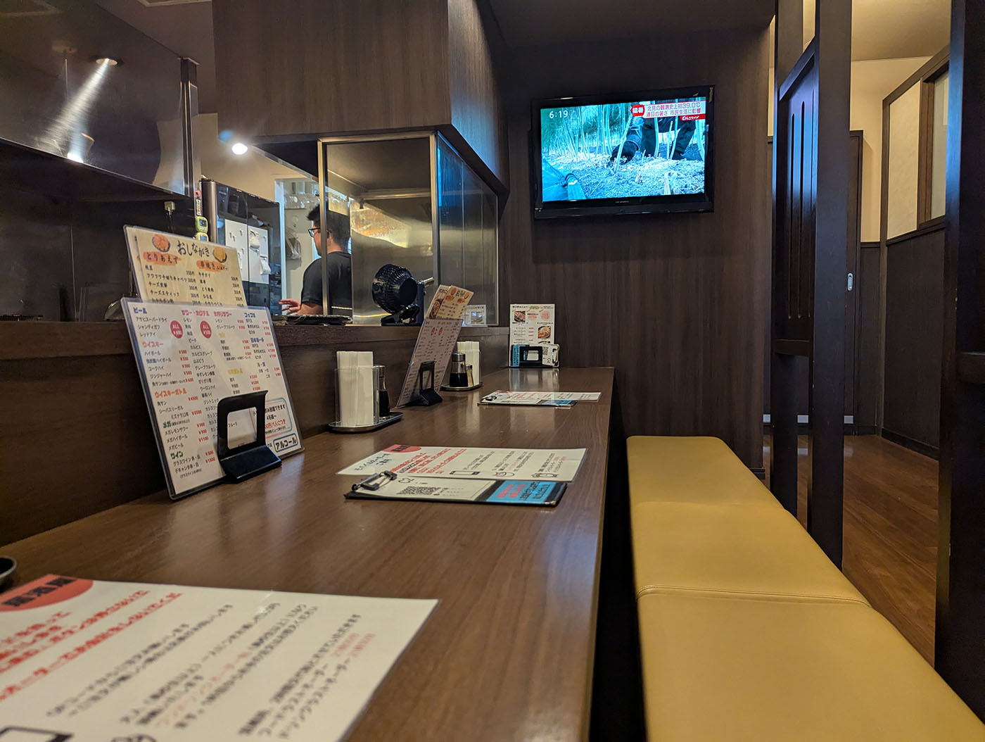 落ち着いた木目調のカウンター席と調理場、奥にはテレビが設置された店内の様子。