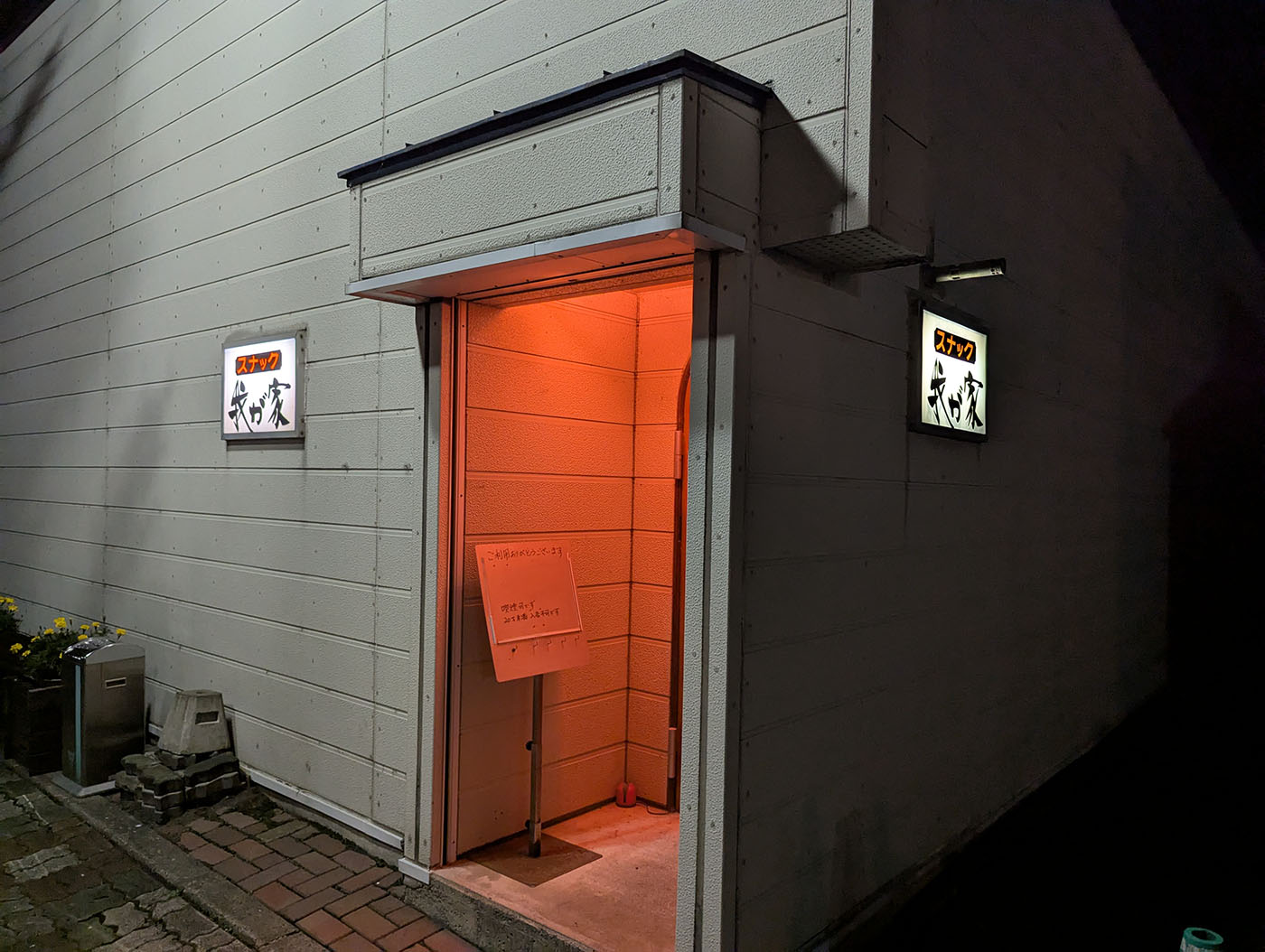 夜の街角に佇む「スナック我が家」の入り口。赤い照明が漏れ、両脇に看板が光っている。