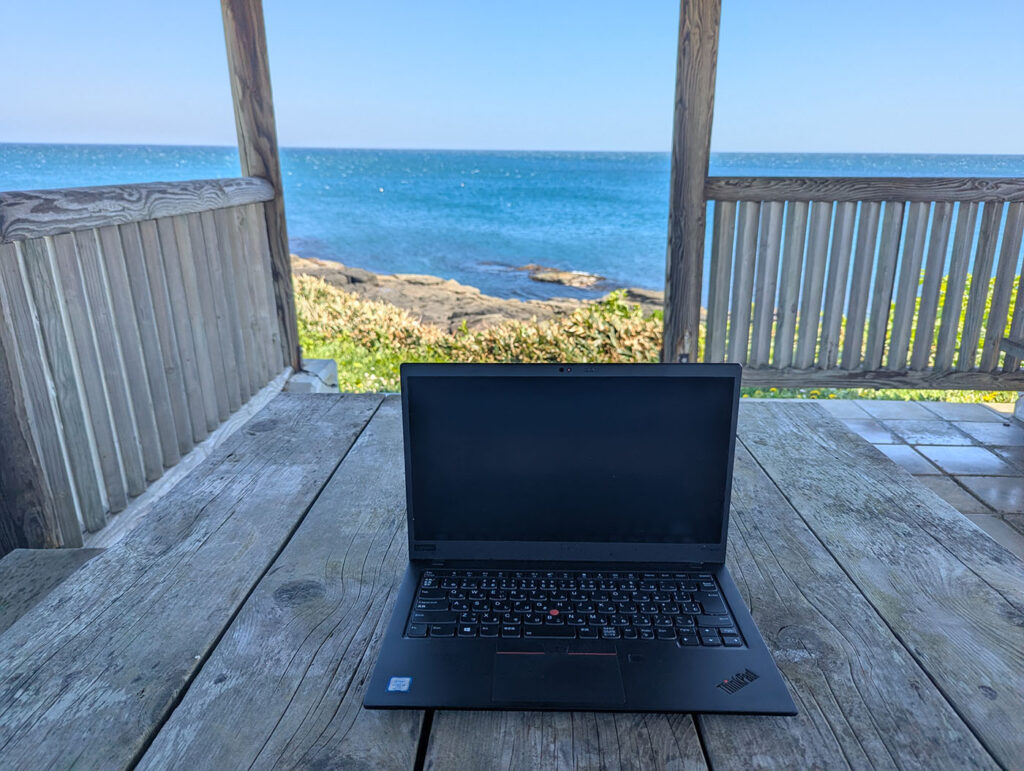 海辺のあずまやにノートPCを置き、青い海を背景に潮風を感じながら仕事をする車中泊ノマドワークの風景