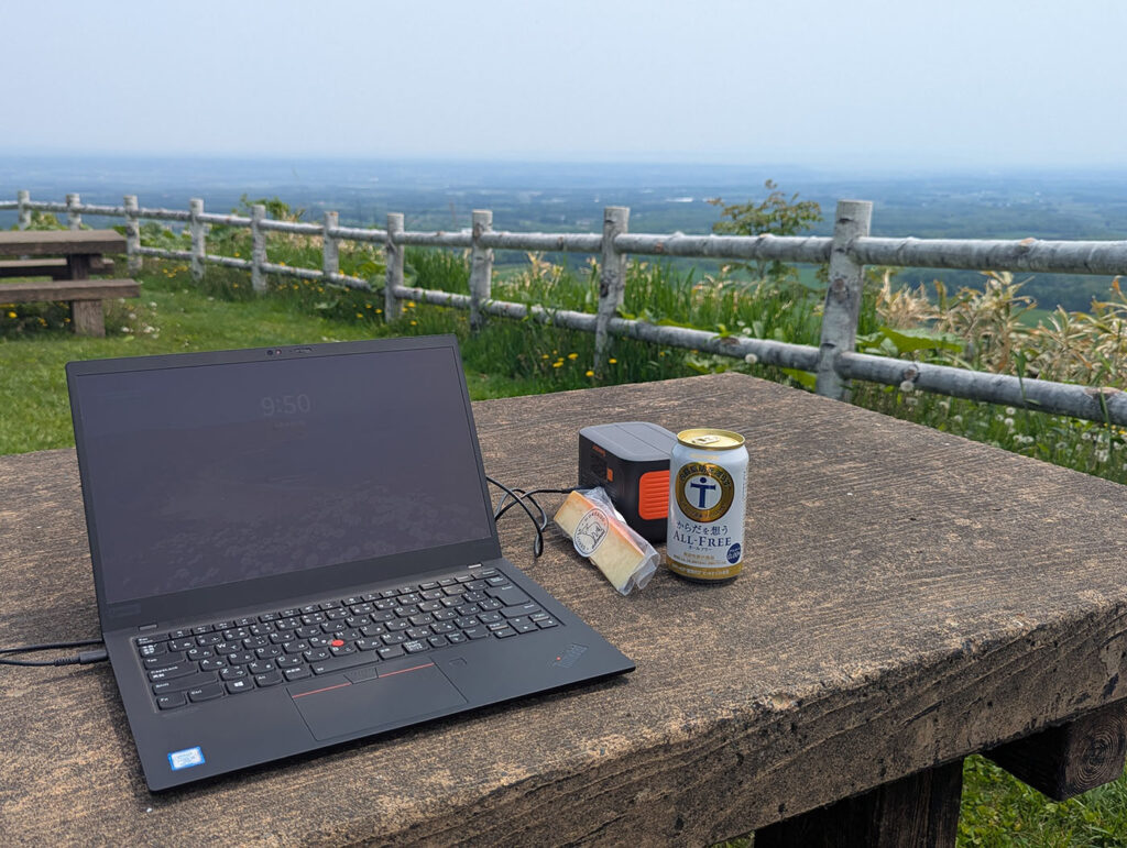 十勝平野を見下ろす高台のベンチでノートPCを広げ、ポータブル電源とノンアルビールを横に置いてノマドワークする様子