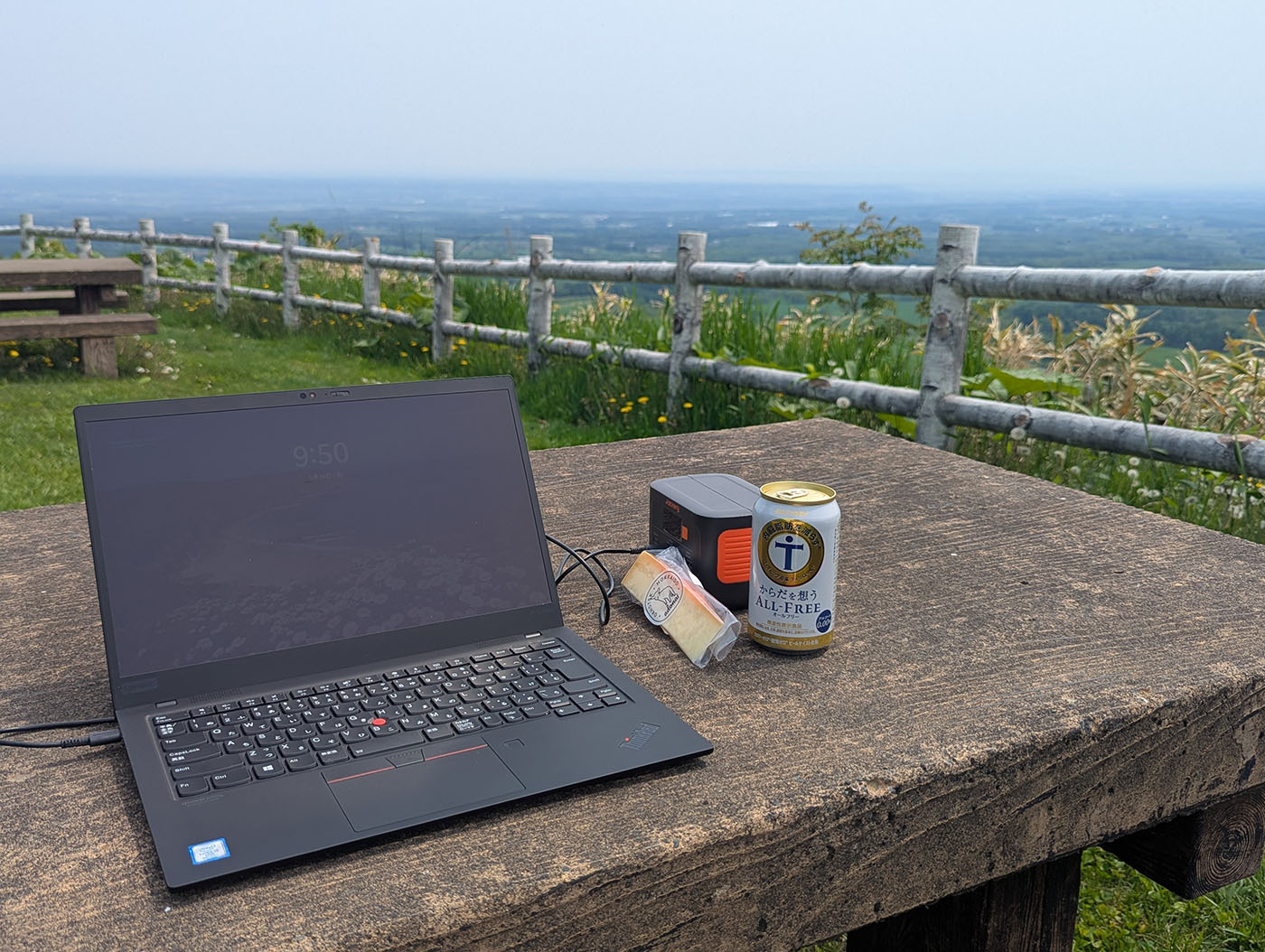 十勝平野を見下ろす高台のベンチでノートPCを広げ、ポータブル電源とノンアルビールを横に置いてノマドワークする様子