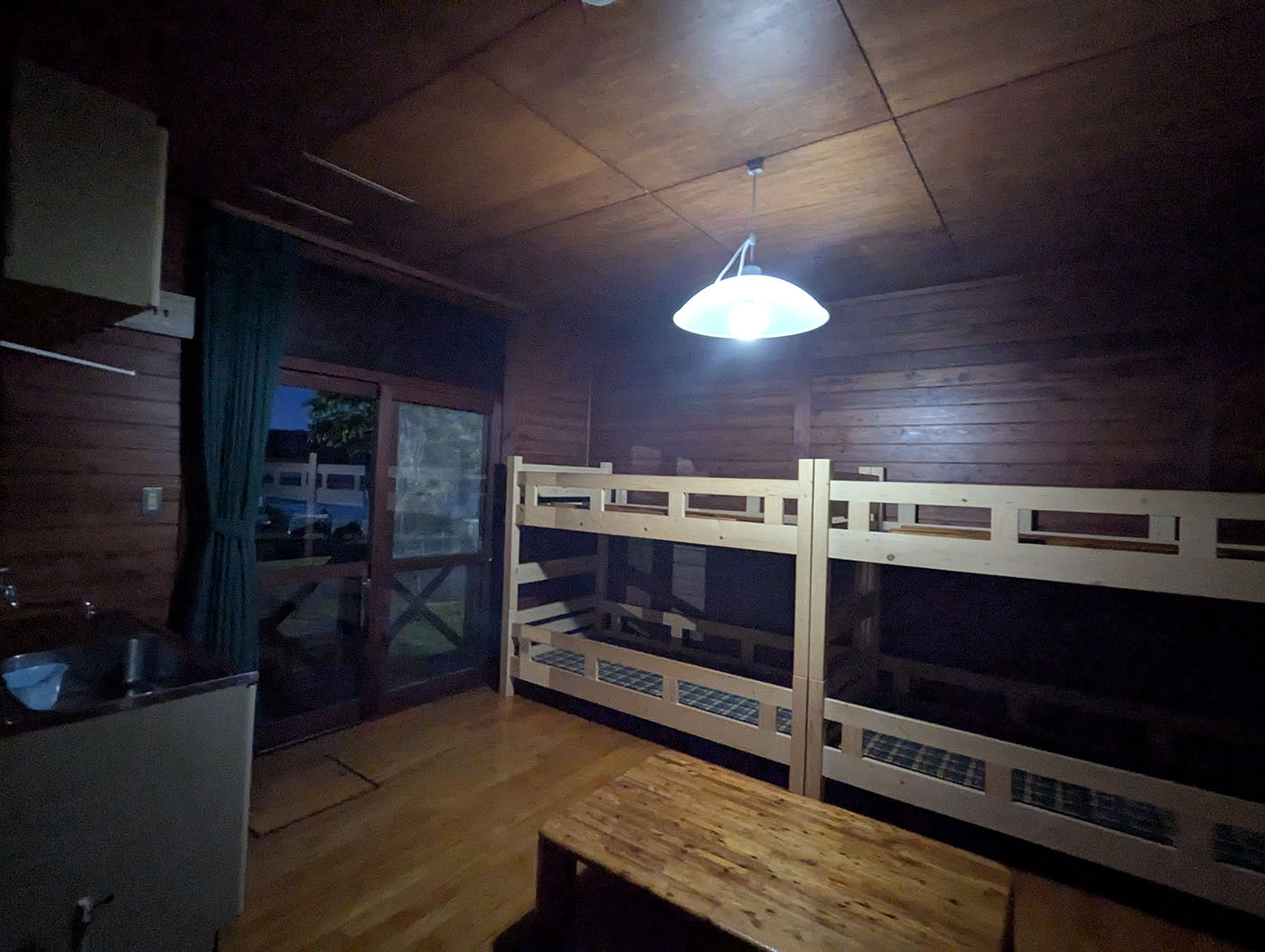 達古武キャンプ場の木の温もりを感じるロッジの室内。二段ベッドと小さな流し台があり、窓の外には夕暮れの景色がのぞく。
