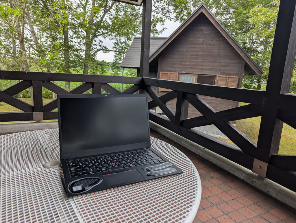 達古武キャンプ場の管理棟テラスにノートPCを置き、森とロッジを背景に早朝のノマドワークを行う様子