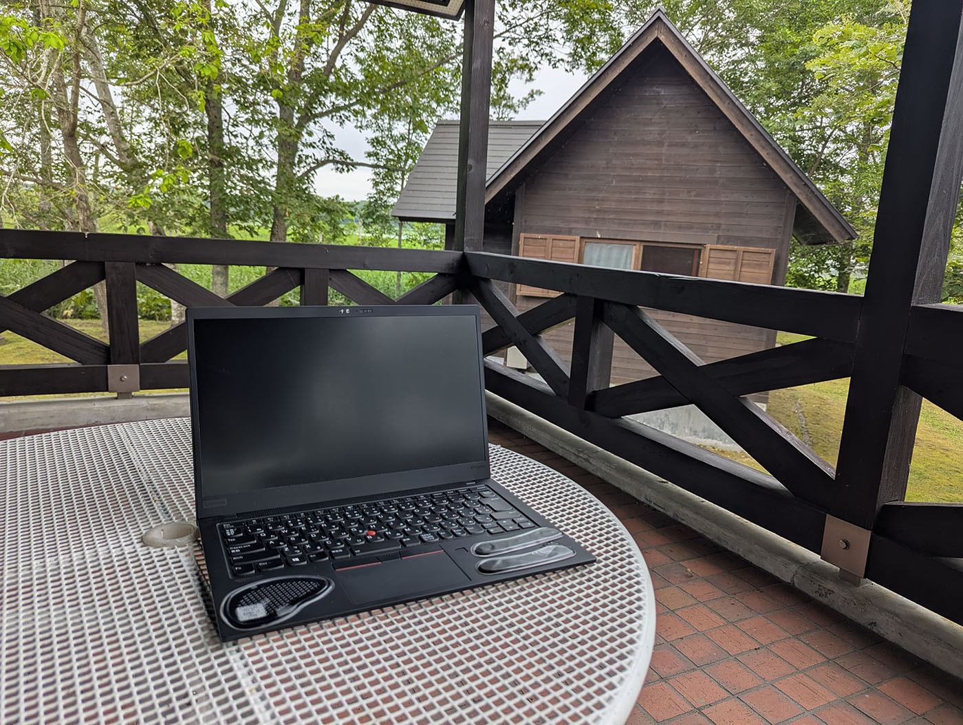 達古武キャンプ場の管理棟テラスにノートPCを置き、森とロッジを背景に早朝のノマドワークを行う様子
