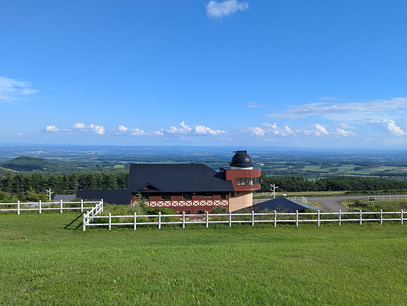 ヌプカの里の丘に建つ天文台付きの建物。手前には白い柵と牧草地が広がり、その向こうに十勝平野の大パノラマが続いている。