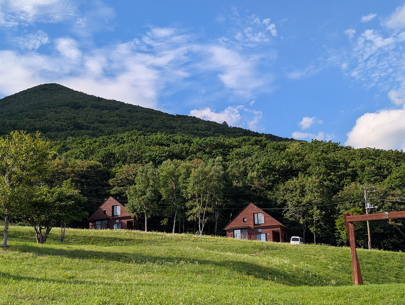 ヌプカの里のコテージ群。緑豊かな山のふもとに赤茶色の三角屋根の建物が並び、芝生の広場と青空が爽やかなコントラストを描いている。